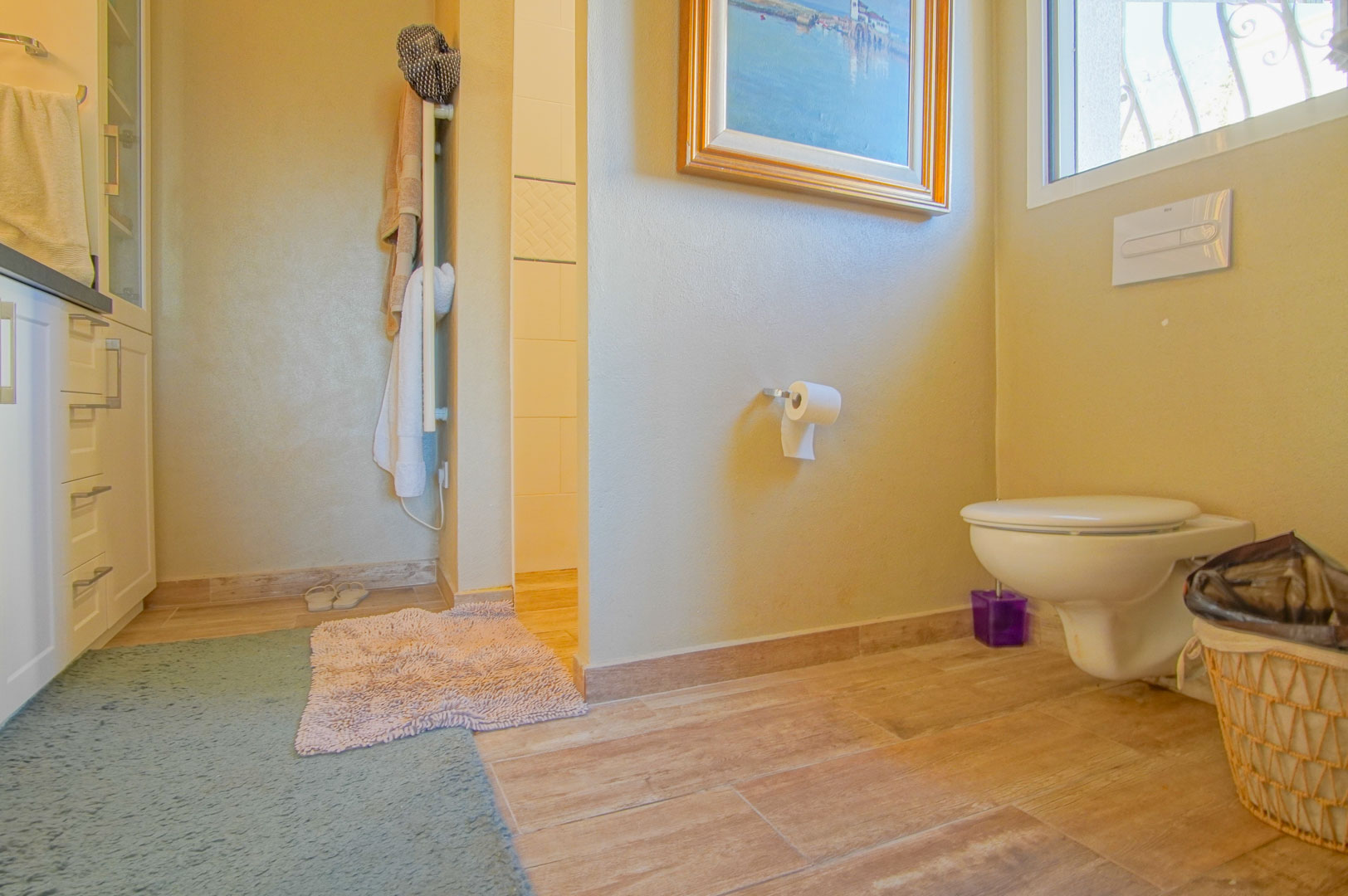 Badkamer met hangend toilet, keramische vloer met houtlook en witte kasten. Voorzien van een handdoekradiator en toegang tot de betegelde inloopdouche.