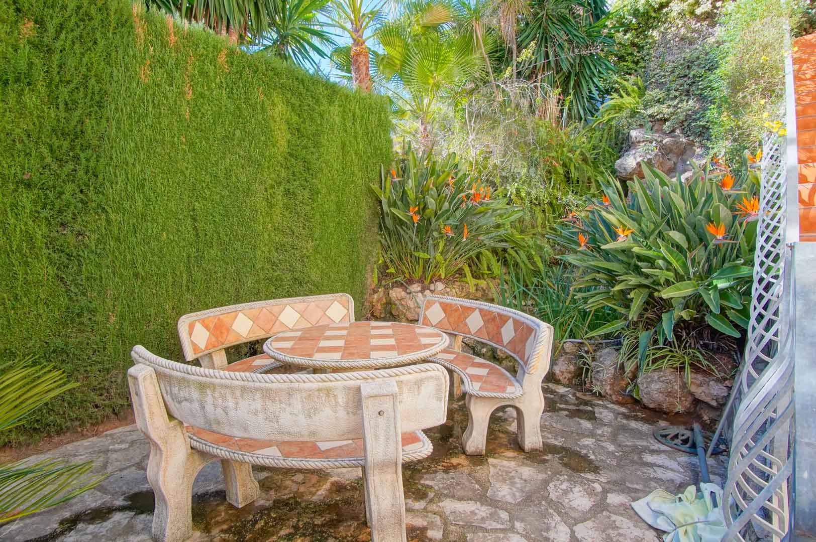 Besloten zithoek buiten met een stenen tafel en gebogen banken met tegelinleg, omgeven door weelderige mediterrane beplanting en paradijsvogelbloemen.
