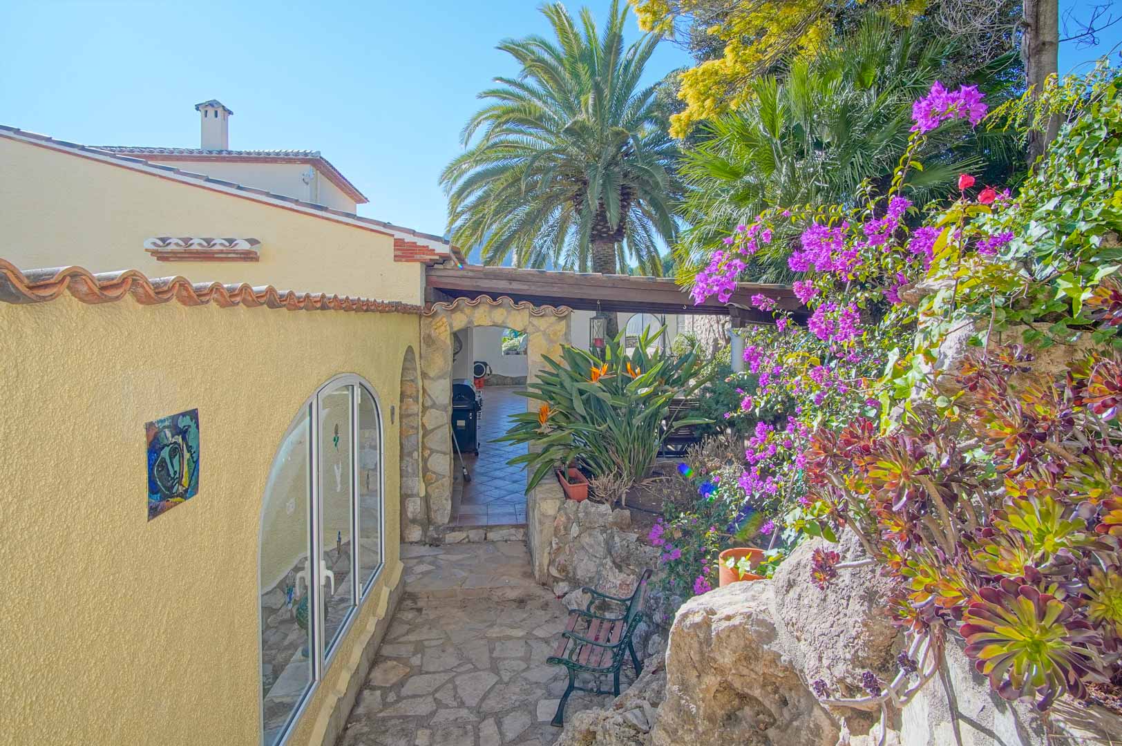 Buitenpad van natuursteen omgeven door bougainville en palmbomen, leidend naar een overdekt terras via een rustieke stenen boog in een mediterrane tuin.