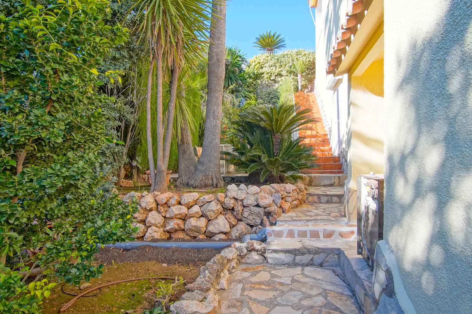 Buitenpad van natuursteen omgeven door stapelmuren en mediterrane beplanting, leidend naar een terracotta trap langs de witte gevel van de villa.