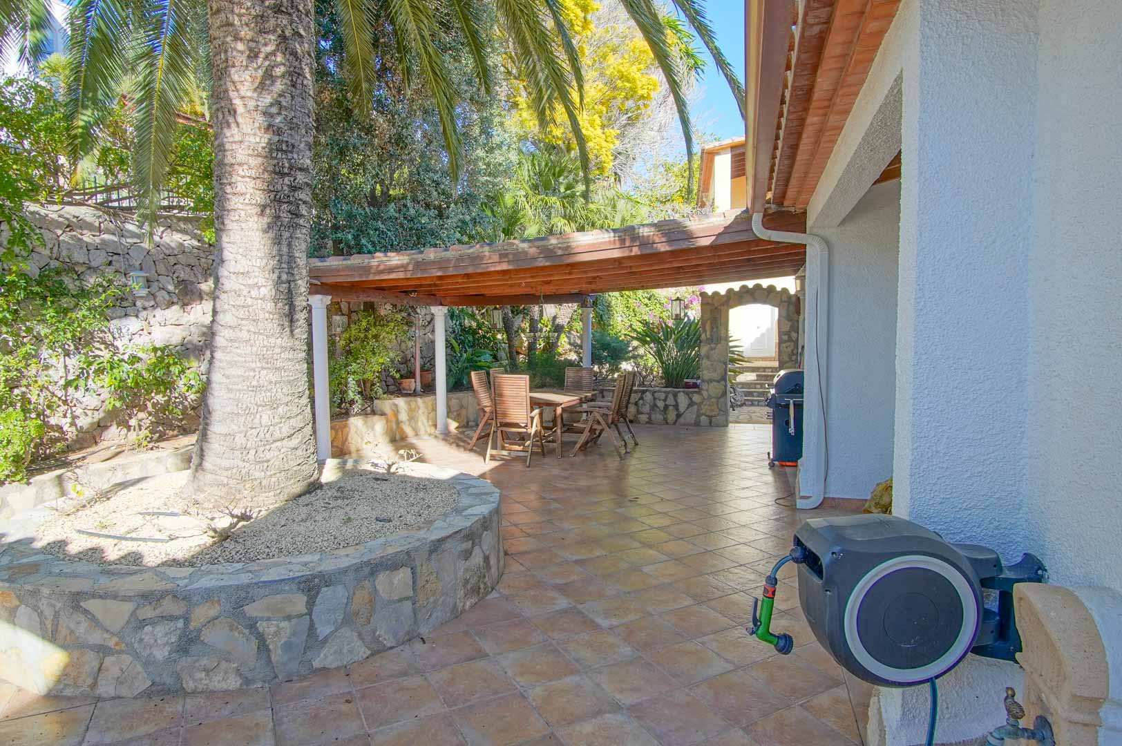 Buitenterras met een houten pergola boven de eethoek, natuurstenen muren en een grote palmboom op een betegelde patio.