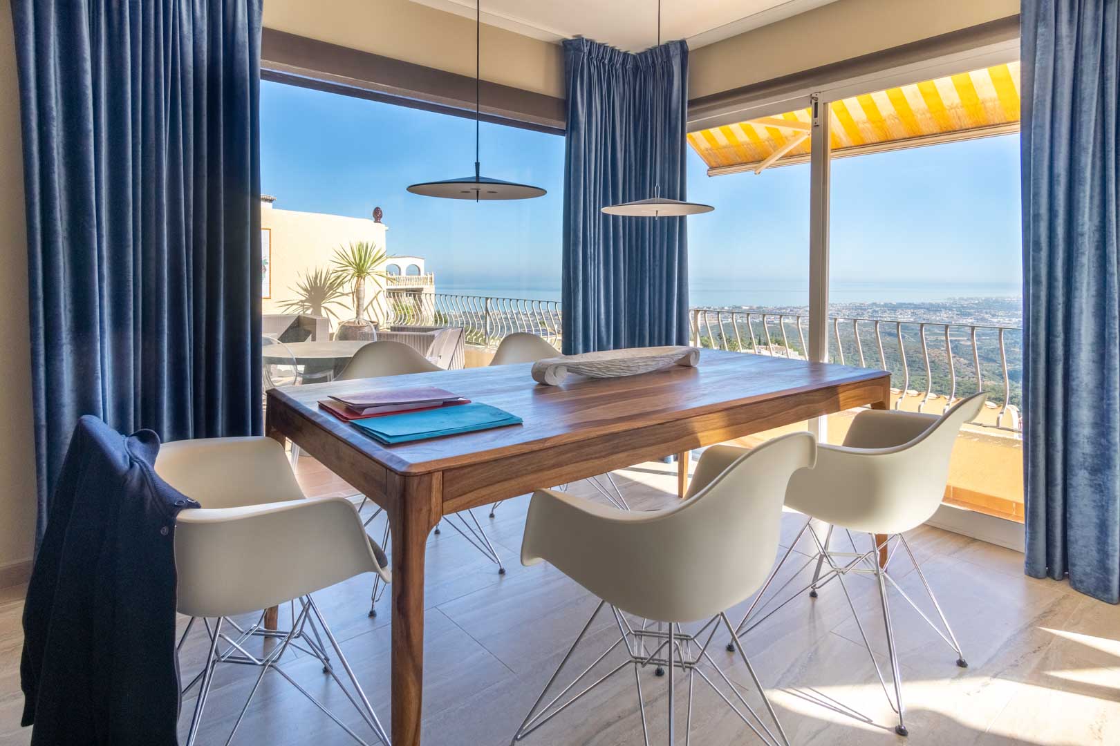 Eethoek met een massief houten tafel en designstoelen, voorzien van kamerhoge ramen met blauwe gordijnen en weids uitzicht over de Middellandse Zee.