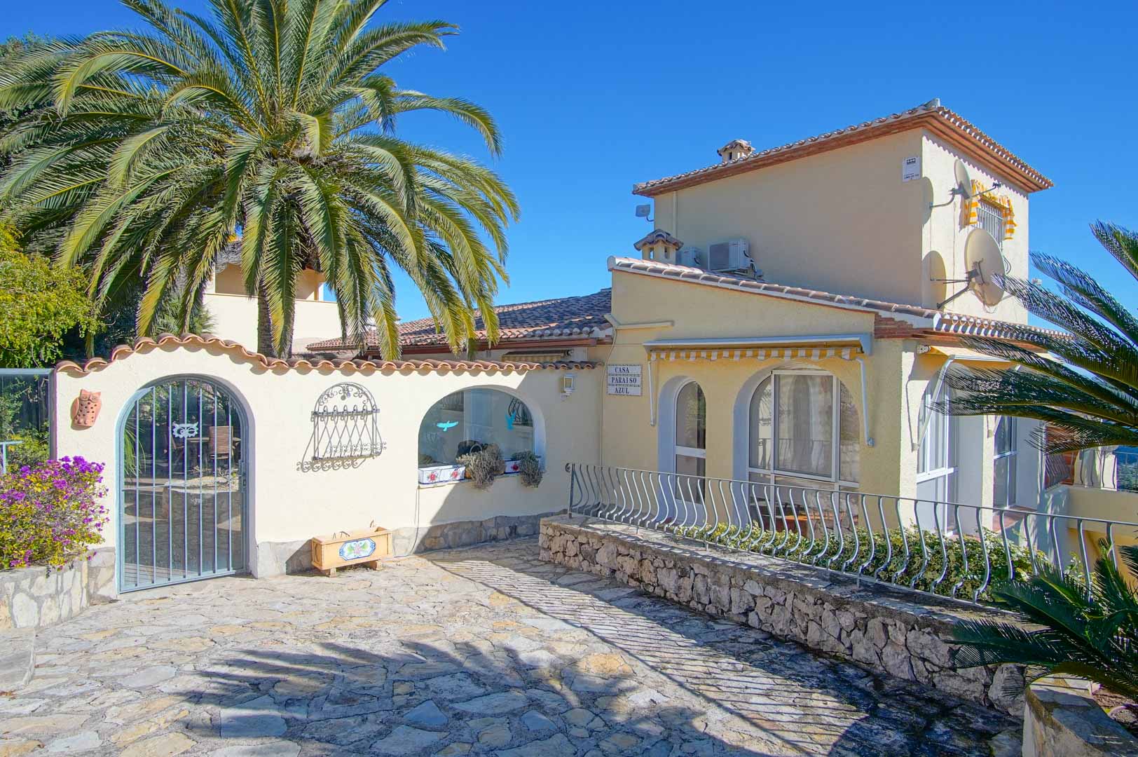 Hoofdgevel van de villa met een met natuursteen geplaveide entree, decoratieve bogen en mediterrane beplanting onder een helderblauwe lucht.