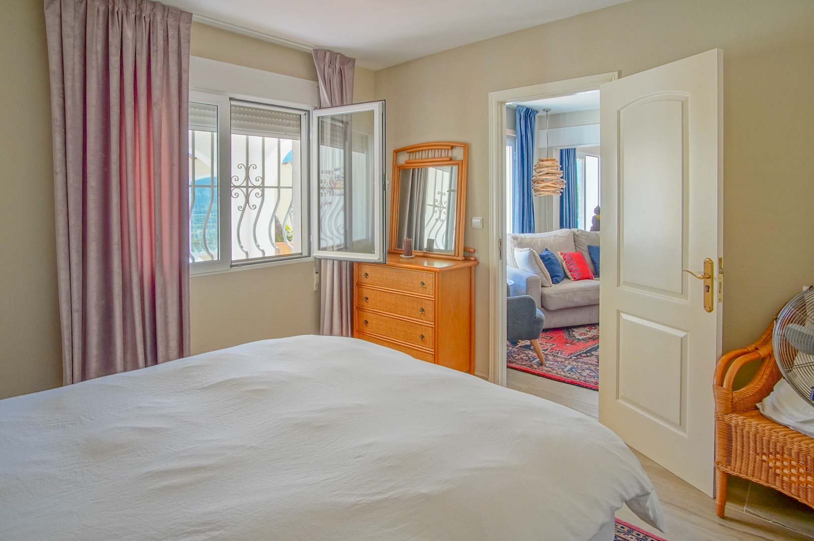Lichte slaapkamer met houten ladekast en raam met decoratieve tralies. Een open deur biedt toegang tot een aangrenzende woonkamer met bank en kleurrijk tapijt.