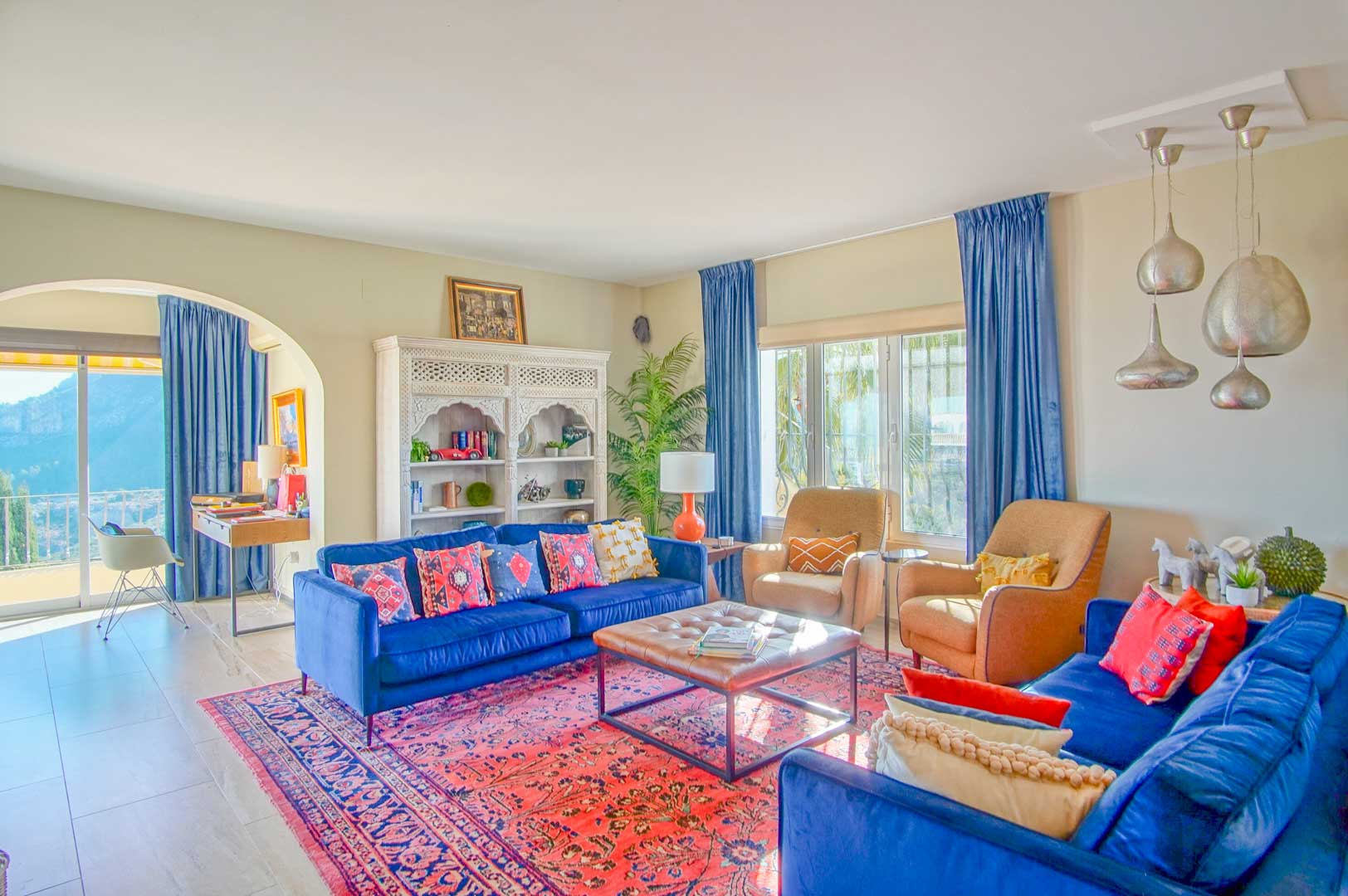 Lichte woonkamer met blauwe fluwelen banken, een oosters tapijt en een wit uitgesneden kast. Een boog leidt naar een werkplek met uitzicht op de bergen.