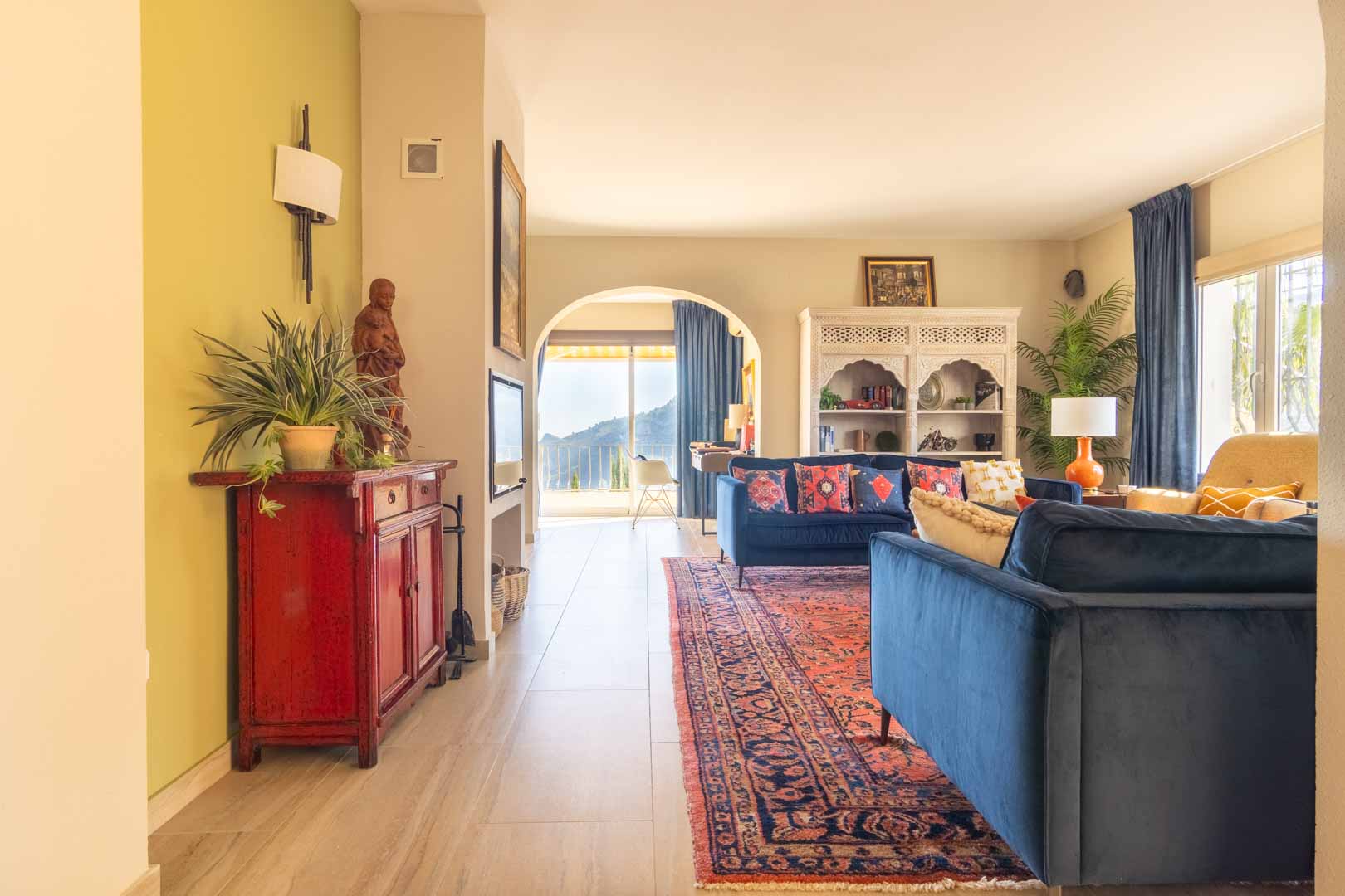 Lichte woonkamer met blauwe fluwelen banken, oosters tapijt en een boogdoorgang met uitzicht op de bergen. Voorzien van bewerkte kasten en een keramische tegelvloer.