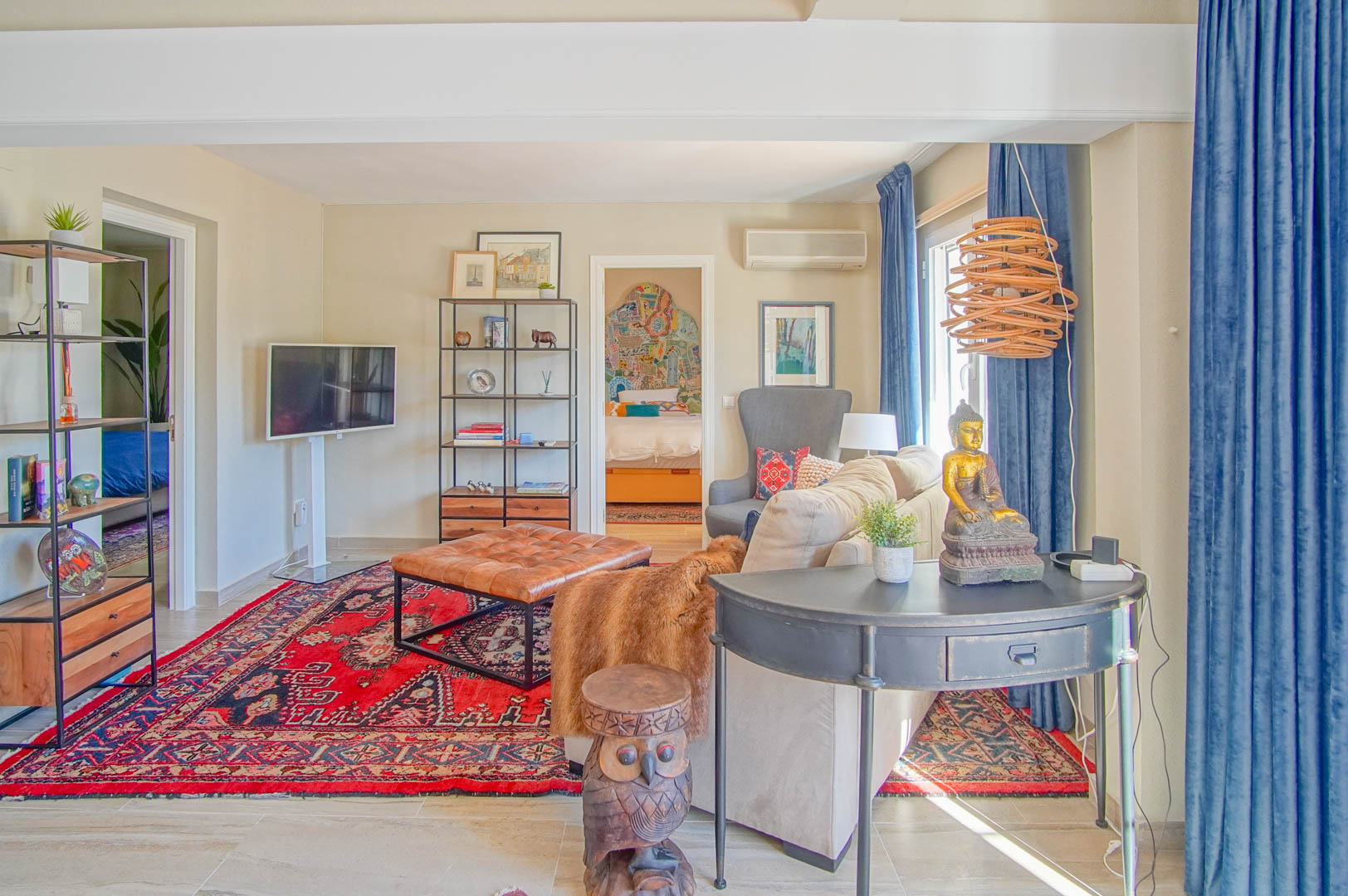 Lichte woonkamer met een rood oosters tapijt, gecapitonneerde lederen hocker en eclectische inrichting, met doorkijk naar een aangrenzende slaapkamer.