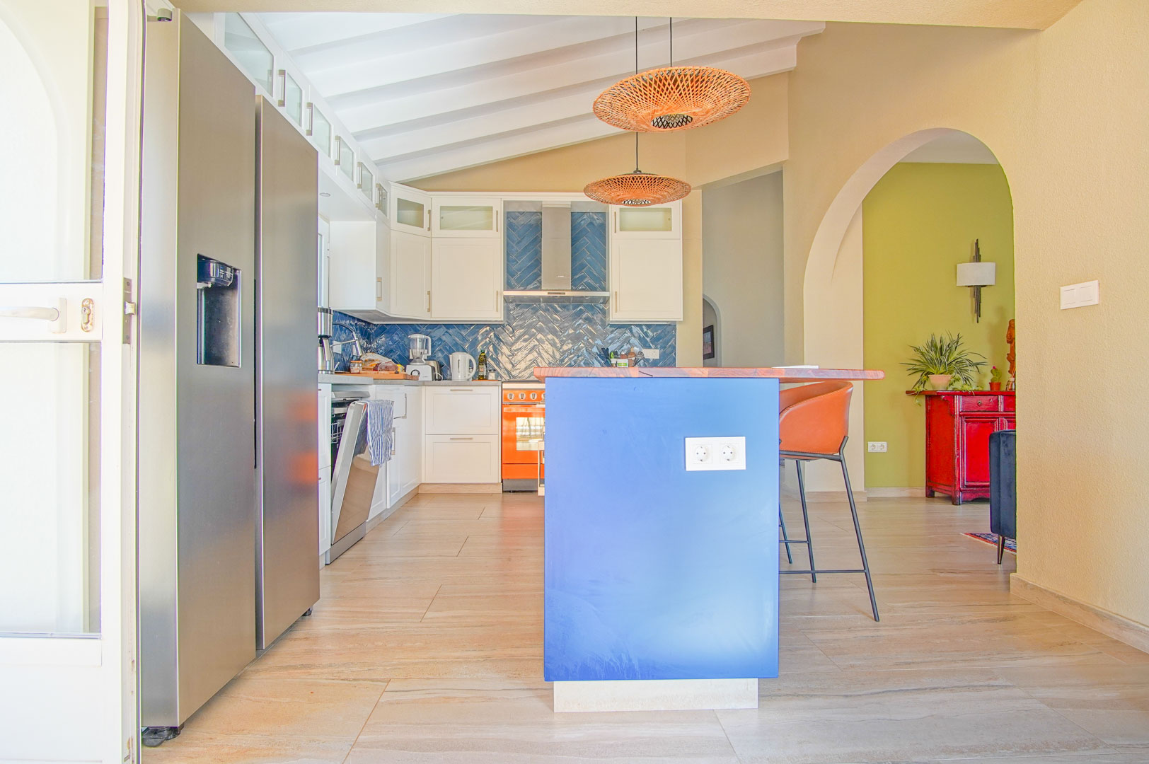 Moderne keuken met blauw kookeiland, houten werkblad en visgraat-tegelwand. Voorzien van een wit balkenplafond, rotan hanglampen en een side-by-side koelkast.
