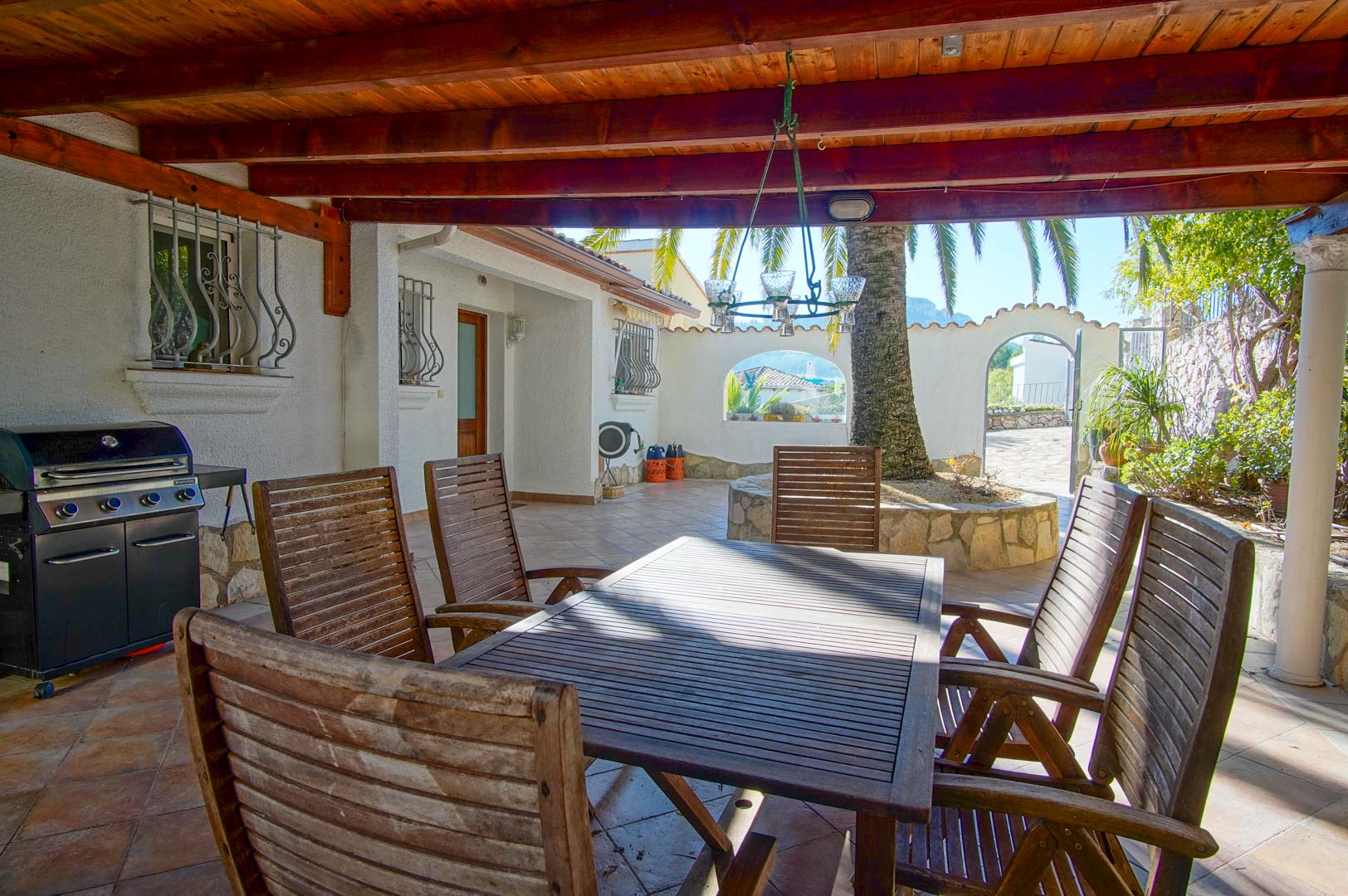 Overdekt terras met houten balkenplafond, buiten-eethoek, gasbarbecue en een binnenplaats met palmboom en natuurstenen muren.