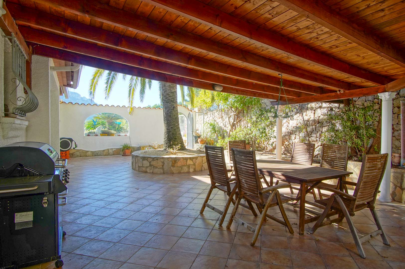 Overdekt terras met houten balkenplafond, eethoek, barbecue en een cirkelvormige stenen plantenbak met palmboom, omgeven door natuurstenen muren.
