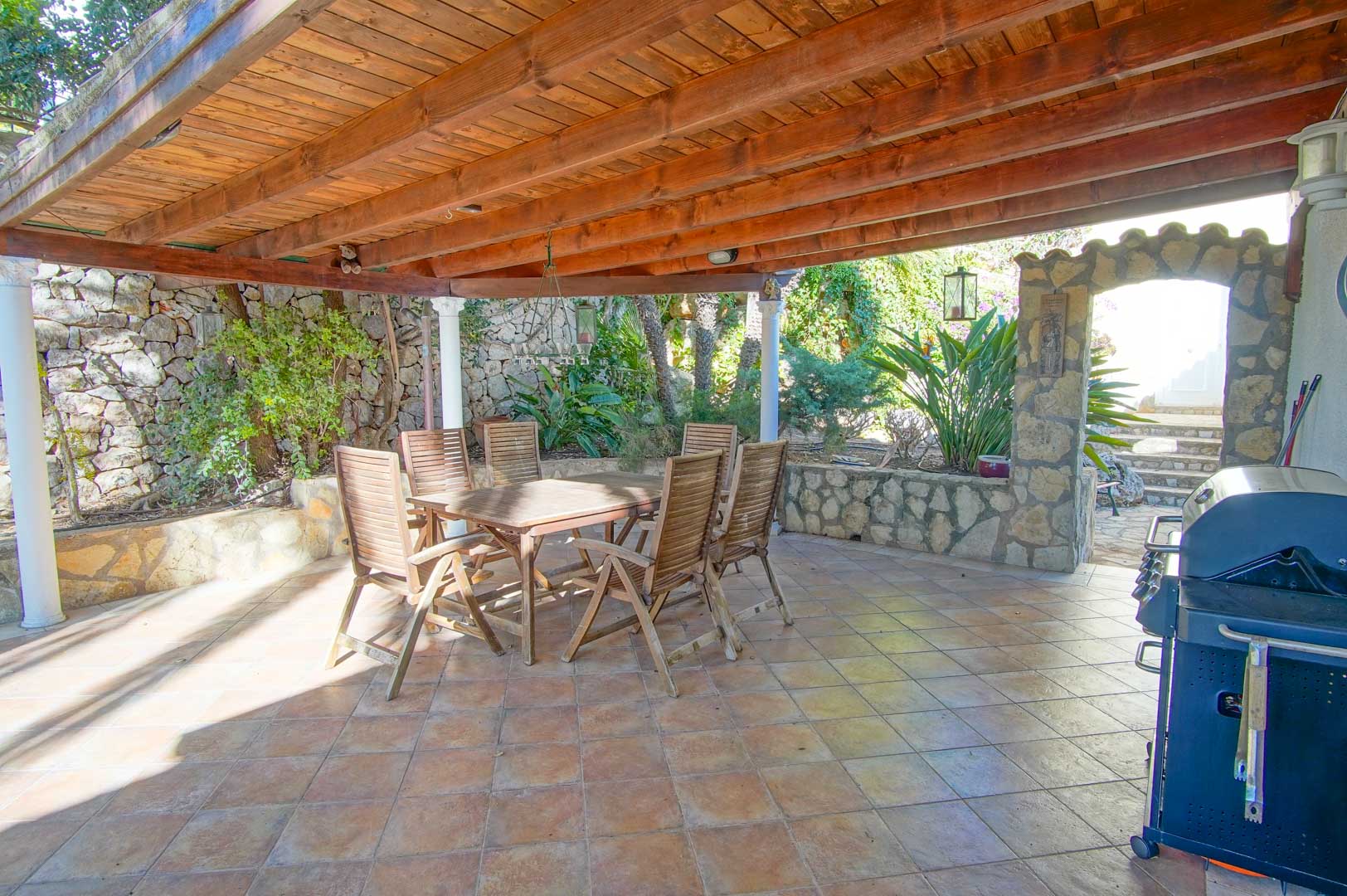 Overdekte buitenruimte met een houten pergola, barbecue, natuurstenen muren en een houten eethoek, omgeven door mediterrane beplanting.
