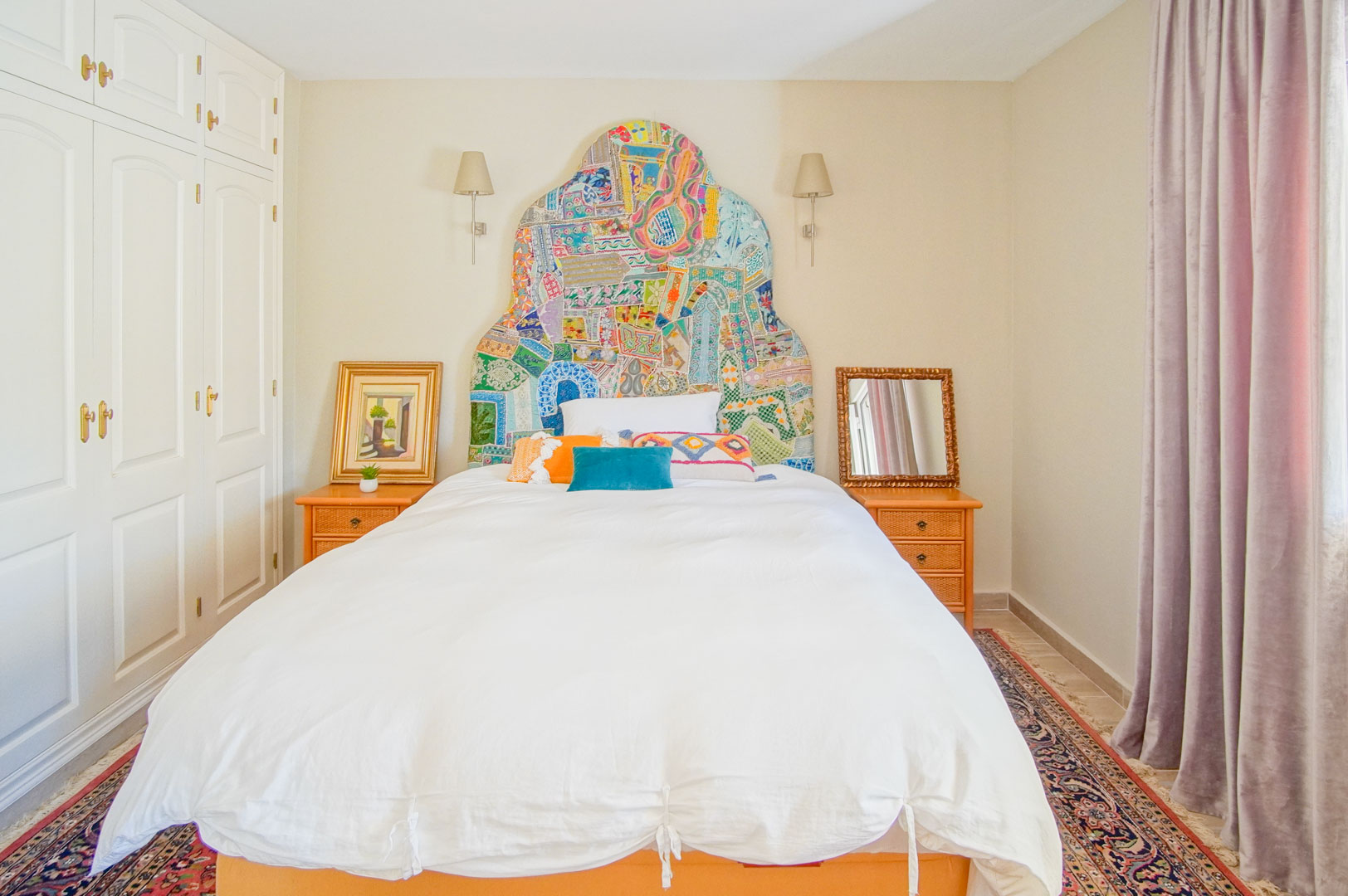 Slaapkamer met een kleurrijk patchwork hoofdbord, witte inbouwkasten en houten nachtkastjes. De ruimte is voorzien van wandverlichting en een oosters tapijt.