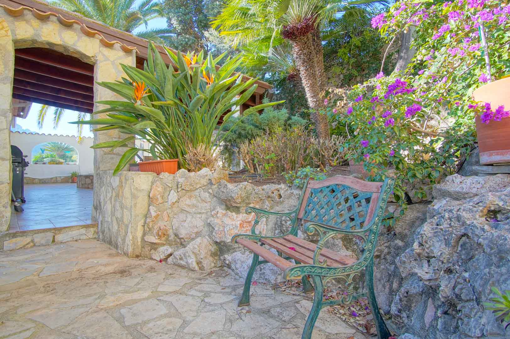 Tuinhoek met een klassieke houten bank, natuurstenen muren en mediterrane beplanting nabij een overdekt terras met authentieke houten balken.