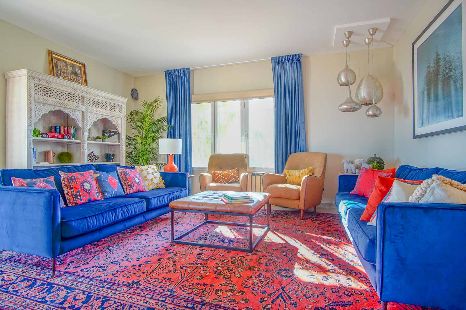 Woonkamer met blauwe fluwelen banken, een rood oosters tapijt en een wit uitgesneden boekenkast. De ruimte heeft veel natuurlijk licht en zilveren hanglampen.