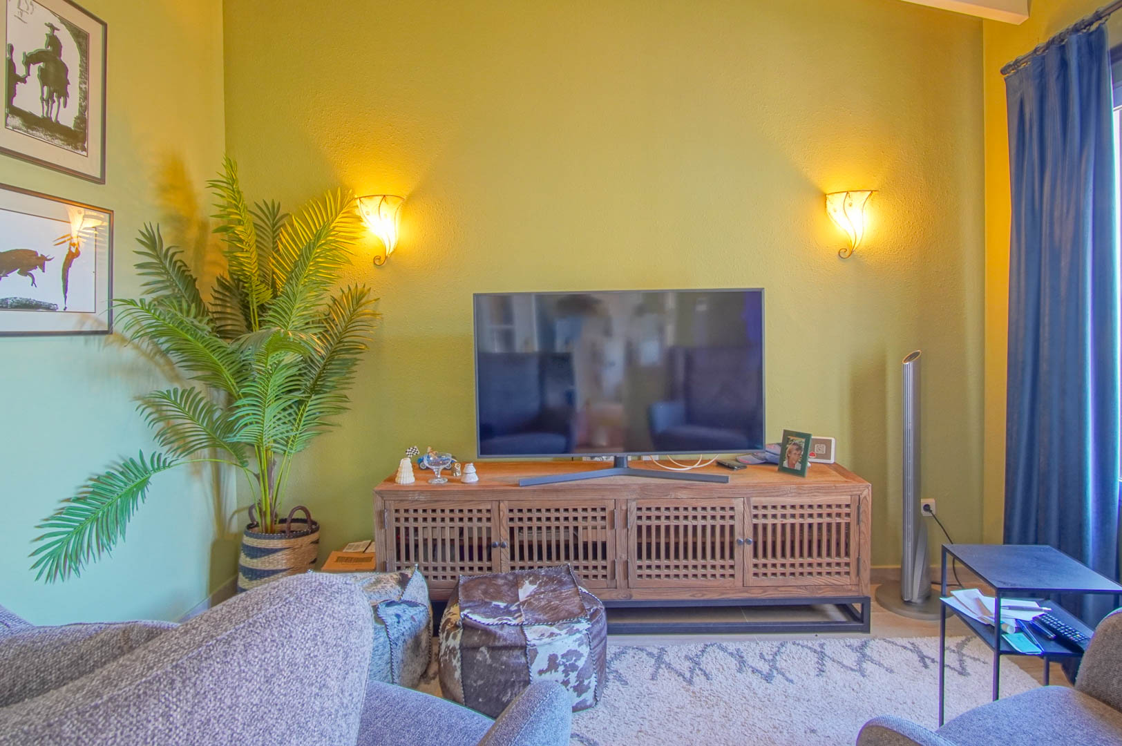 Zithoek met een houten tv-meubel met roosterdeuren, sfeervolle wandverlichting, een grote kamerplant en comfortabele poefs op een vloerkleed.