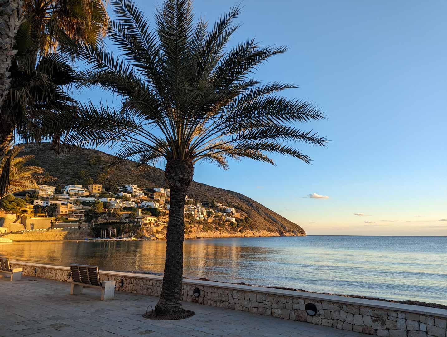 Geplaveide kustpromenade in Moraira met palmbomen en houten banken, uitkijkend over de rustige Middellandse Zee en de villawijk tegen de heuvel bij zonsondergang.
