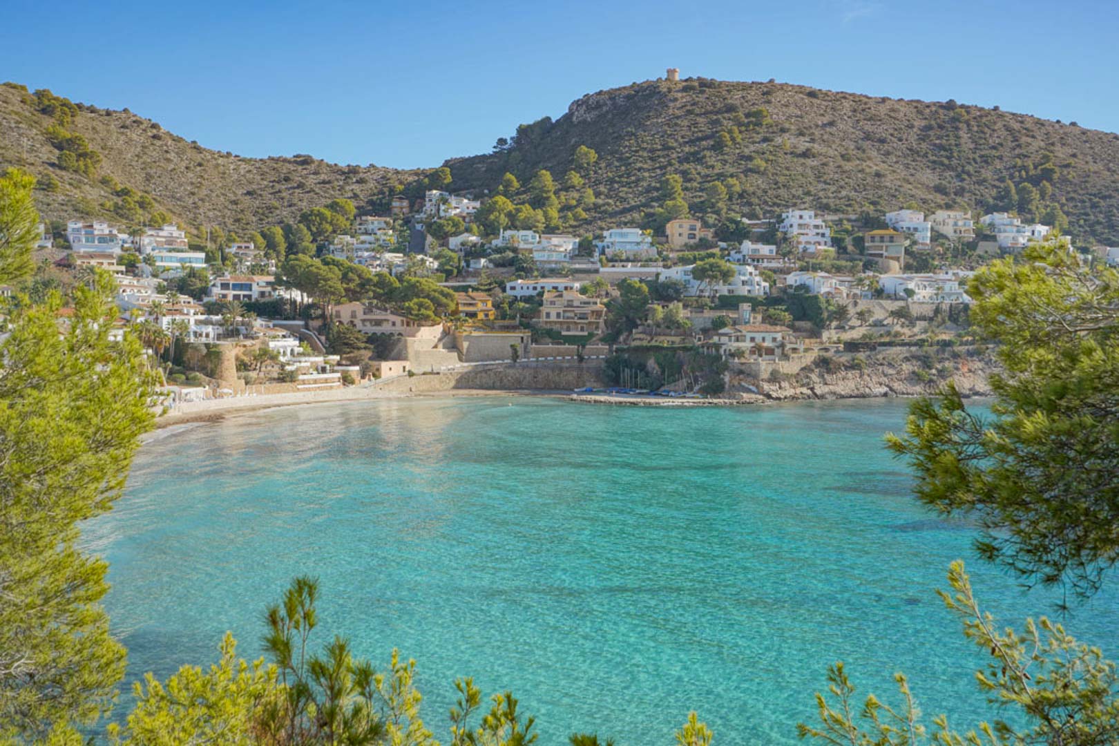 Panoramisch uitzicht op de baai van El Portet in Moraira, met kristalhelder turquoise water, een zandstrand en pijnbomen langs de mediterrane kustlijn.