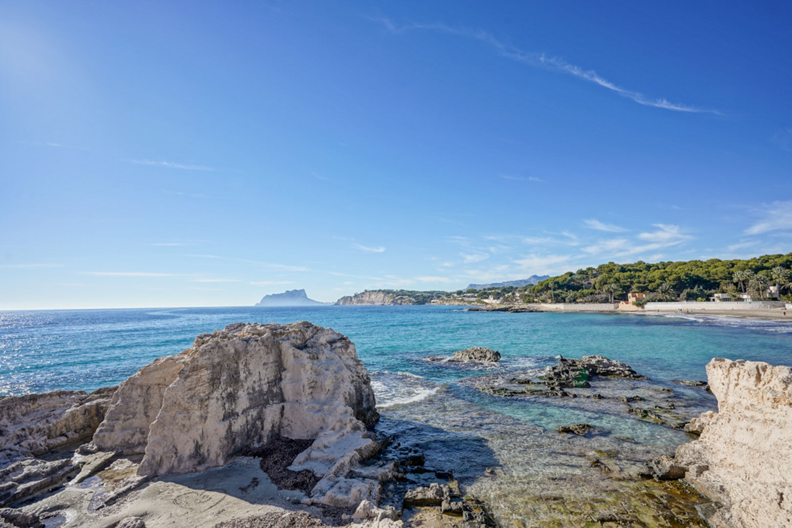 Panoramisch uitzicht op de kustlijn van Moraira met natuurlijke rotsformaties, kristalhelder turquoise water en de Peñón de Ifach aan de horizon.