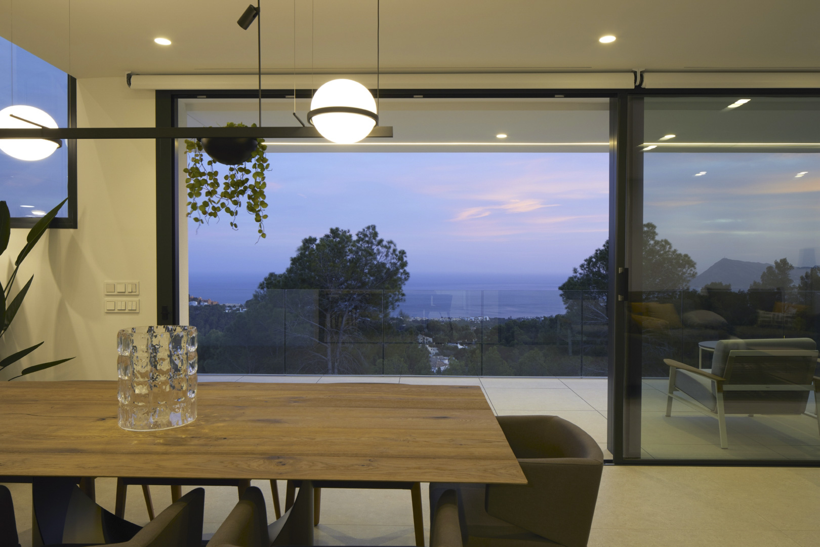 Eetkamer met een robuuste houten tafel en designverlichting, grenzend aan een terras met glazen balustrades en weids uitzicht over de Middellandse Zee.