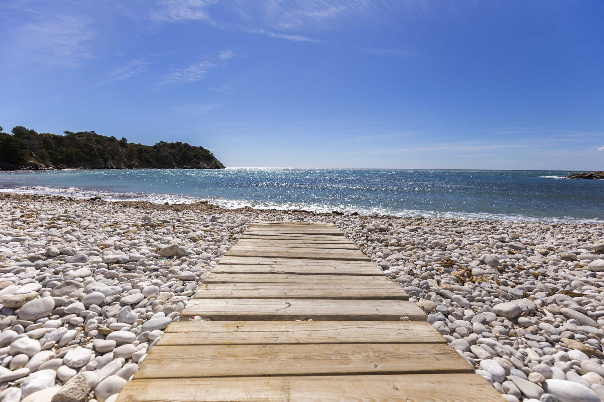 Houten looppad over een wit kiezelstrand met uitzicht op de turquoise Middellandse Zee en de rotsachtige kustlijn van Altea.