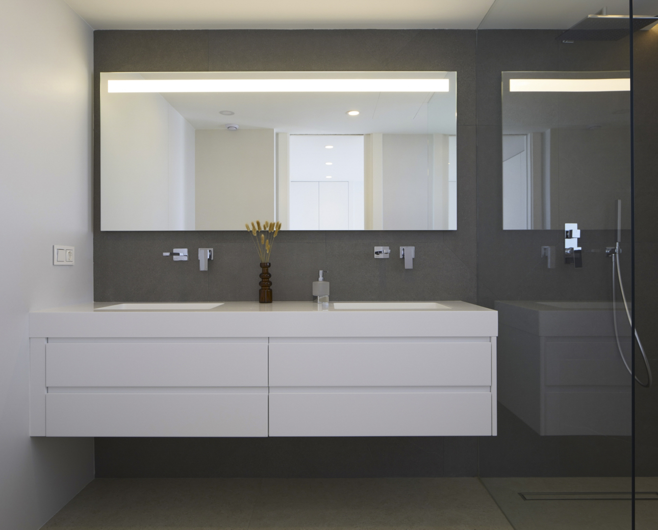 Moderne badkamer met een zwevend wit dubbel wastafelmeubel, inbouwkranen en een grote spiegel met geïntegreerde verlichting tegen een donkere wand.