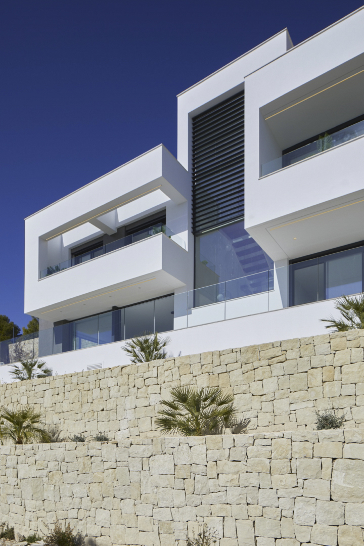 Moderne gevel met witte geometrische volumes en glazen balustrades, gelegen boven een natuurstenen stapelmuur met jonge palmbomen.