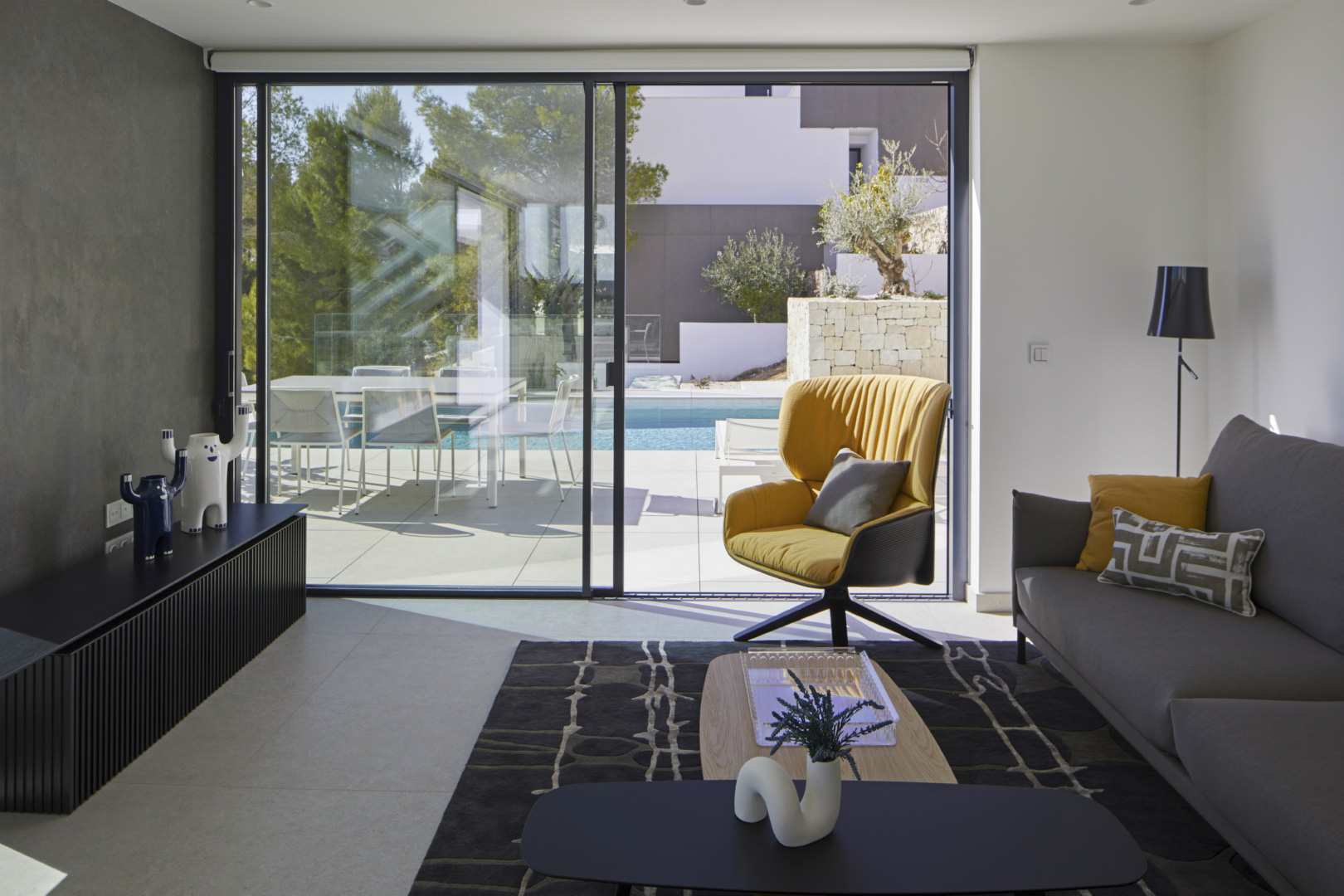 Moderne woonkamer met grote glazen schuifdeuren die toegang bieden tot het terras en het zwembad. Voorzien van designmeubels, een gele fauteuil en een tegelvloer.