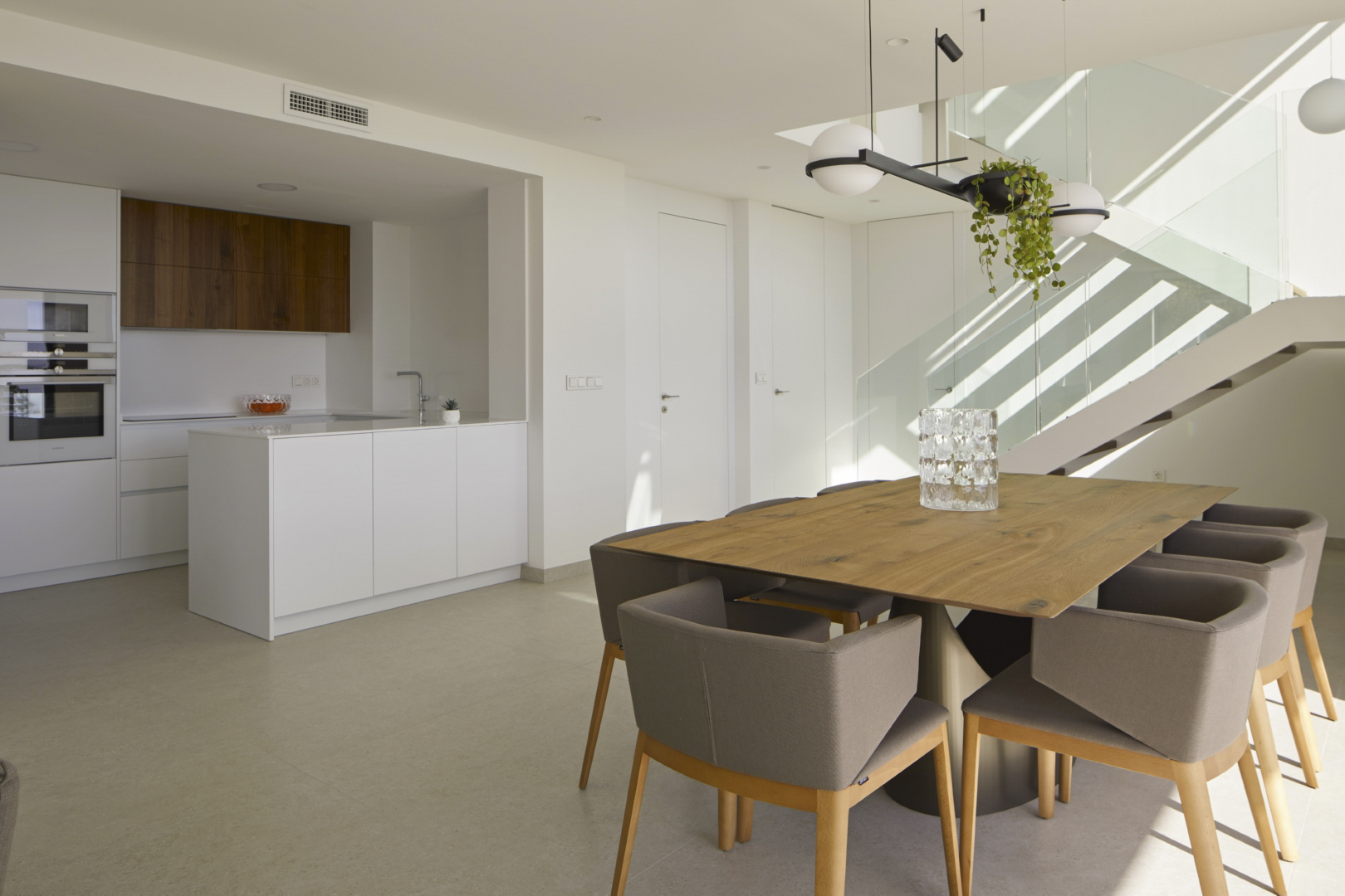 Open eetkamer met houten tafel, minimalistische keuken met wit kookeiland en een moderne trap met glazen balustrade.