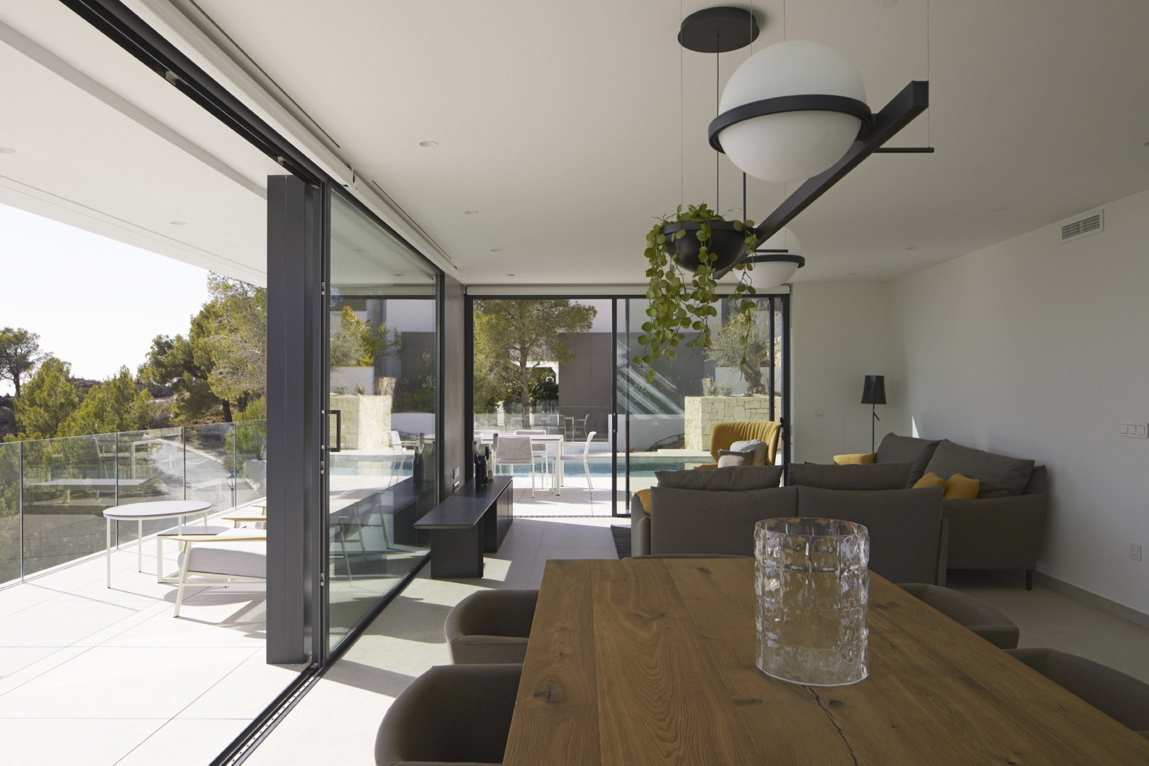 Open woon- en eetkamer met massief houten tafel, design hanglampen en grote glazen schuifdeuren die toegang bieden tot het terras en het overloopzwembad.