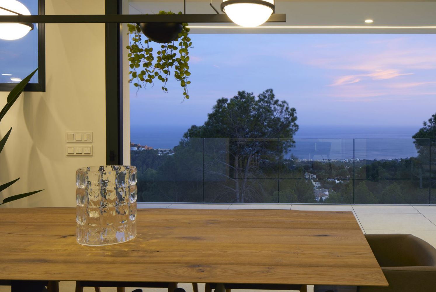 Uitzicht vanuit de eethoek met massief houten tafel. Grote glazen schuifdeuren bieden toegang tot het terras met glazen balustrade en panoramisch zeezicht bij zonsondergang.