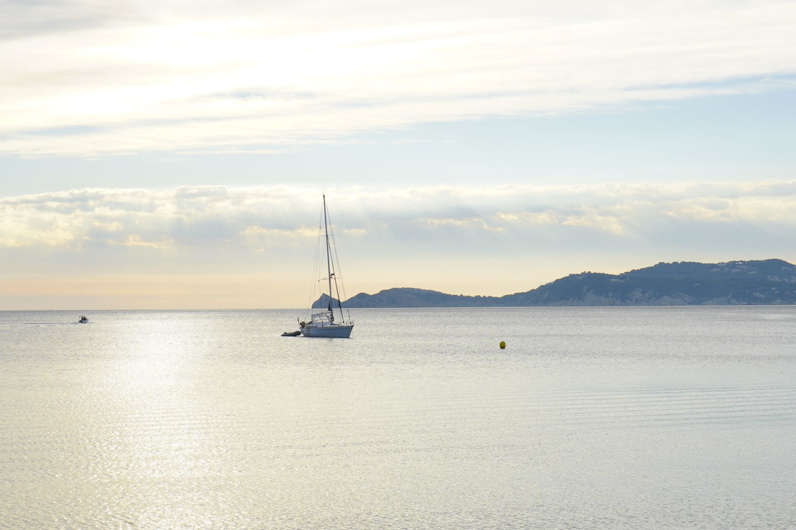 Panoramisch uitzicht op de Middellandse Zee in Jávea met een zeilboot op kalm water en zacht licht dat door de wolken op de kustlijn valt.