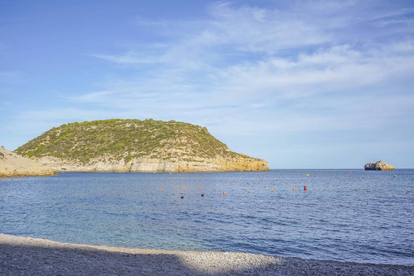 Panoramisch uitzicht op een kiezelstrand in Jávea, met kristalhelder mediterraan water en omliggende kustkliffen onder een heldere blauwe lucht.