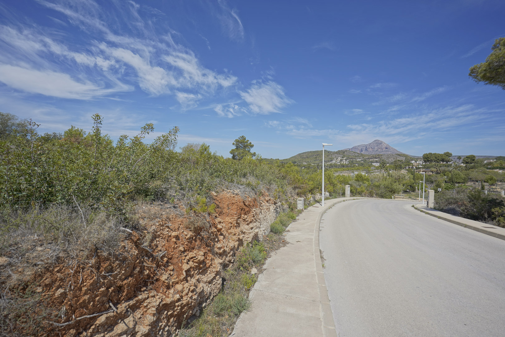 Uitzicht op de toegangsweg in de woonwijk Valsol in Jávea, met de berg Montgó op de achtergrond en inheemse mediterrane beplanting.