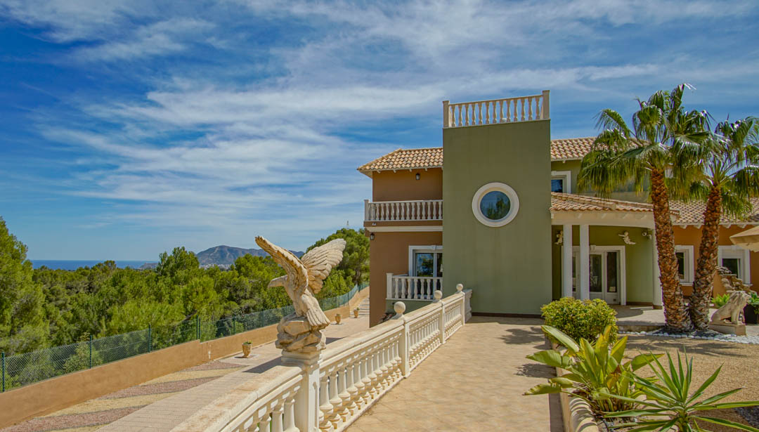 Exterieurgevel met klassieke balustrades, een decoratief adelaarsbeeld en een entree met palmbomen die uitkijkt over het mediterrane dennenbos en de zee.