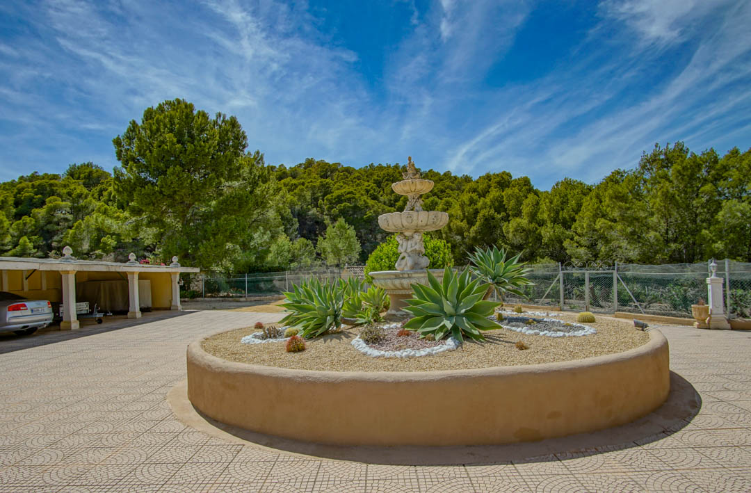 Hoofdingang met een klassieke fontein met drie niveaus en een ronde plantenbak met agaves, naast een overdekte carport en dennenbos.