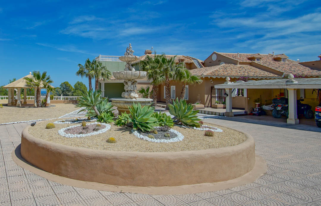 Hoofdingang met een sierlijke stenen fontein, een ronde plantenbak met agaves en cactussen, een overdekte carport en een tuinpaviljoen.