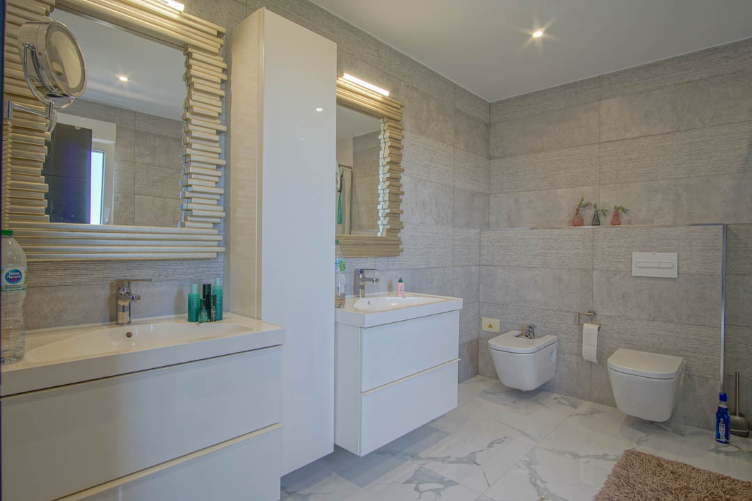 Moderne badkamer met dubbele wastafel, witte zwevende meubels en spiegels met houten lamellenlijsten. Afgewerkt met grijze wandtegels en marmeren vloer.