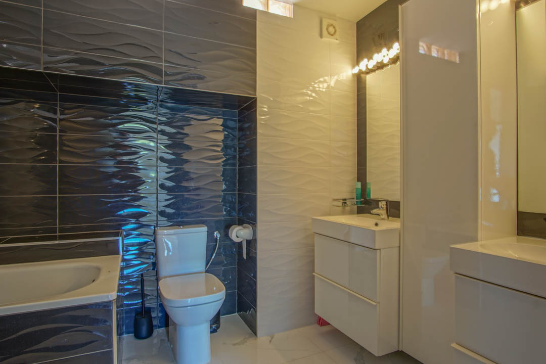 Moderne badkamer met getextureerde golfpatroon tegels in wit en donkergrijs, een strak wit wastafelmeubel en geïntegreerde verlichting boven de spiegel.