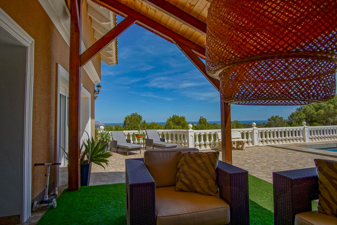Overdekt terras met houten balken en zithoek, met panoramisch uitzicht op de Middellandse Zee achter een klassieke witte balustrade.