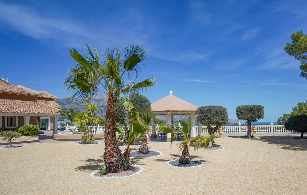 Ruime grindtuin met palmbomen en vormsnoei struiken, voorzien van een witgekolomd paviljoen met panoramisch uitzicht op de zee en de bergen.