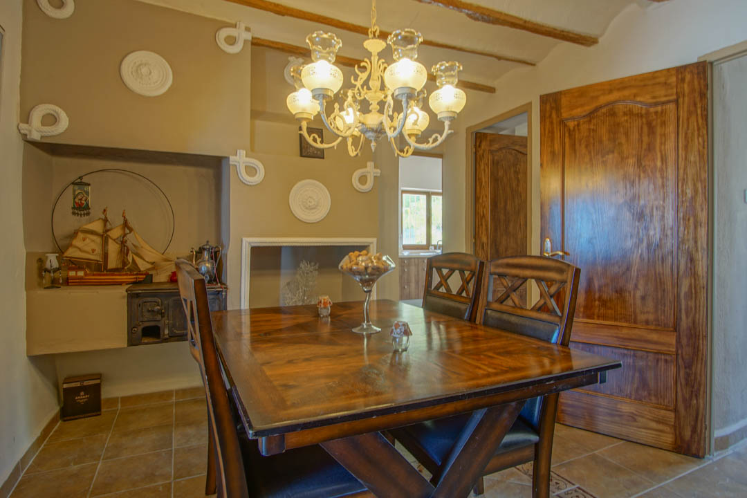 Rustieke eetkamer met een massief houten tafel, zichtbare plafondbalken en een klassieke kroonluchter. Een traditionele nis bevat een antieke kachel en decoratieve elementen.