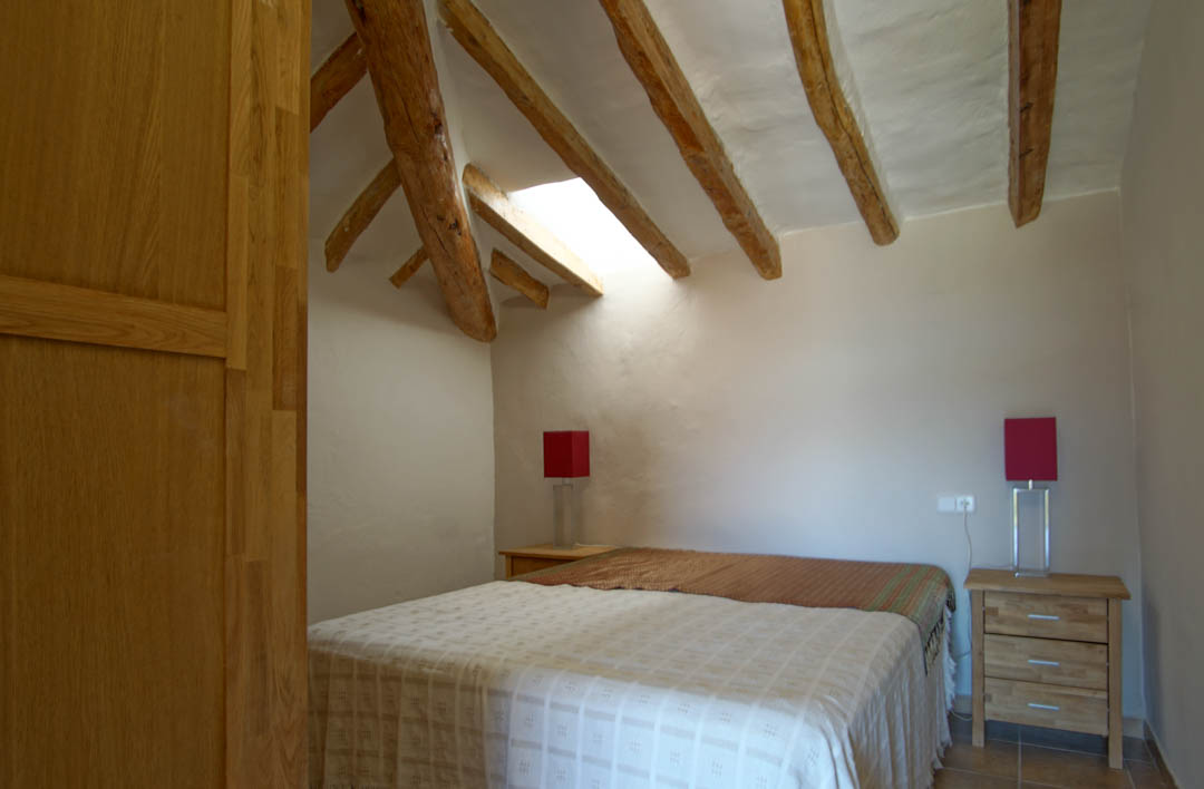 Rustieke slaapkamer met authentieke houten balken, een dakraam voor natuurlijk licht en witte muren. Voorzien van houten nachtkastjes en rode accentlampen.