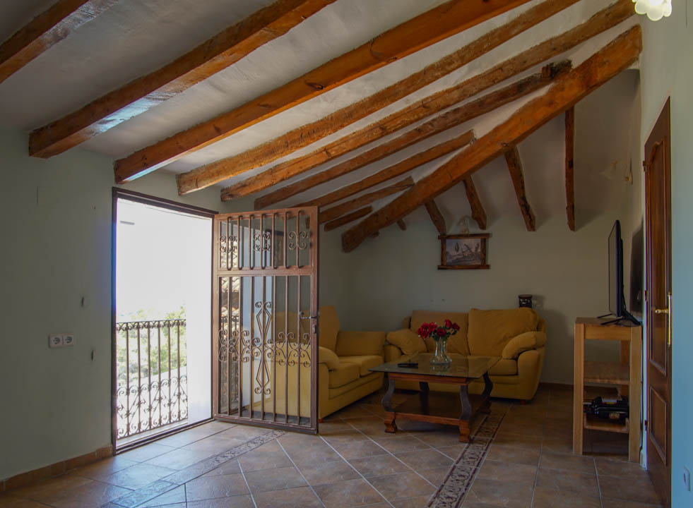 Rustieke woonkamer met authentieke houten plafondbalken, tegelvloer met sierrand en toegang tot het balkon via een sierlijk smeedijzeren hekwerk.