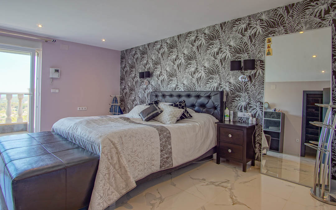 Slaapkamer met marmerlook vloer, tropisch behang op de accentmuur, gecapitonneerd lederen hoofdbord en toegang tot een balkon.