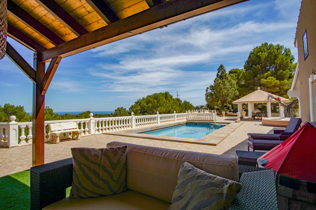 Terras met zwembad, witte balustrade en panoramisch zeezicht. Voorzien van een zithoek onder een houten pergola en een paviljoen tussen mediterrane pijnbomen.
