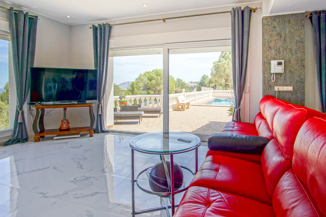 Woonkamer met wit marmeren vloer, rood lederen bank en grote glazen schuifdeuren die toegang bieden tot het terras met zwembad en panoramisch uitzicht.
