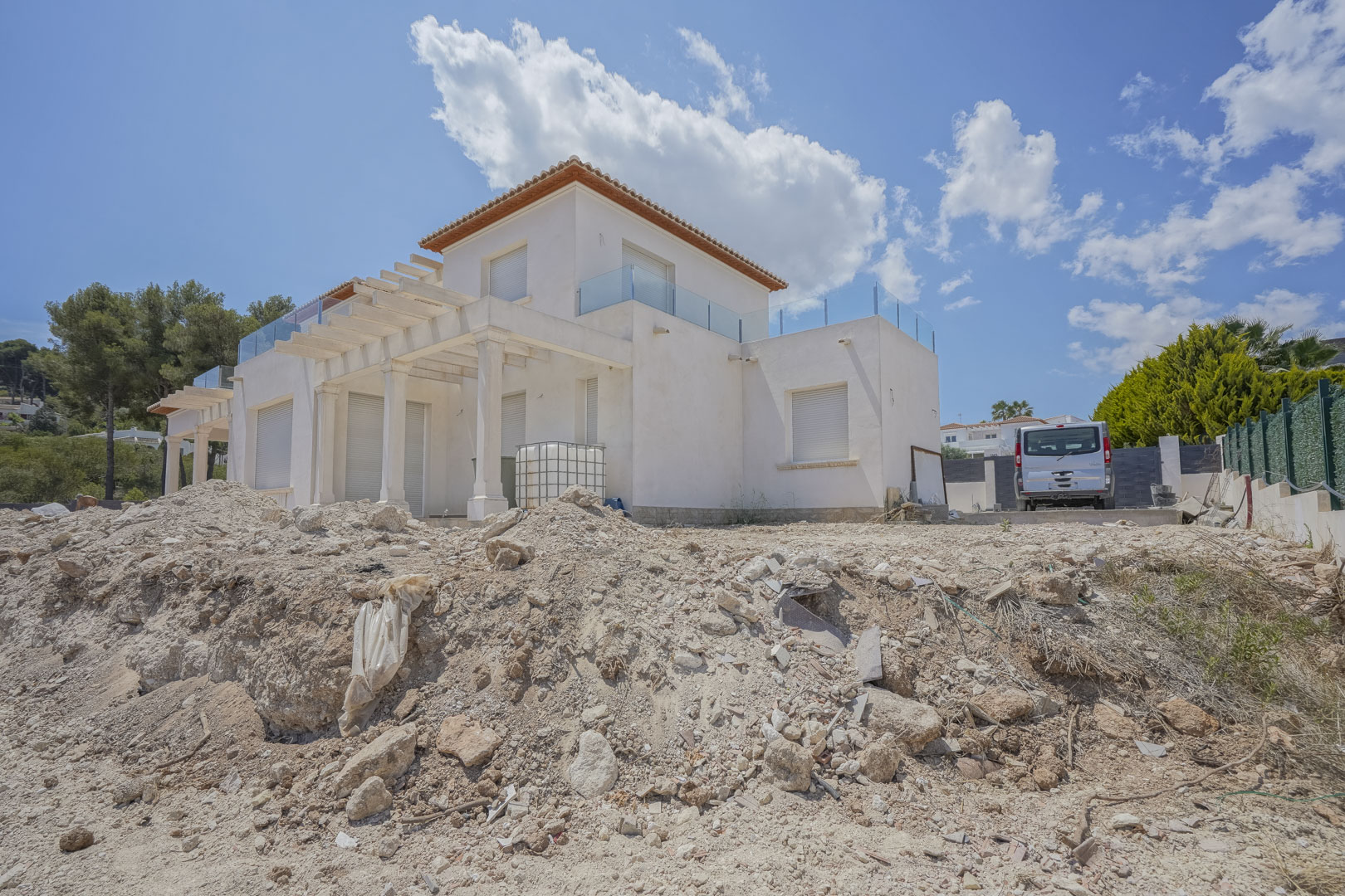 Buitenaanzicht van de villa in de laatste bouwfase, met witte gevels, pergola's met kolommen en terrassen op de bovenverdieping met glazen balustrades.