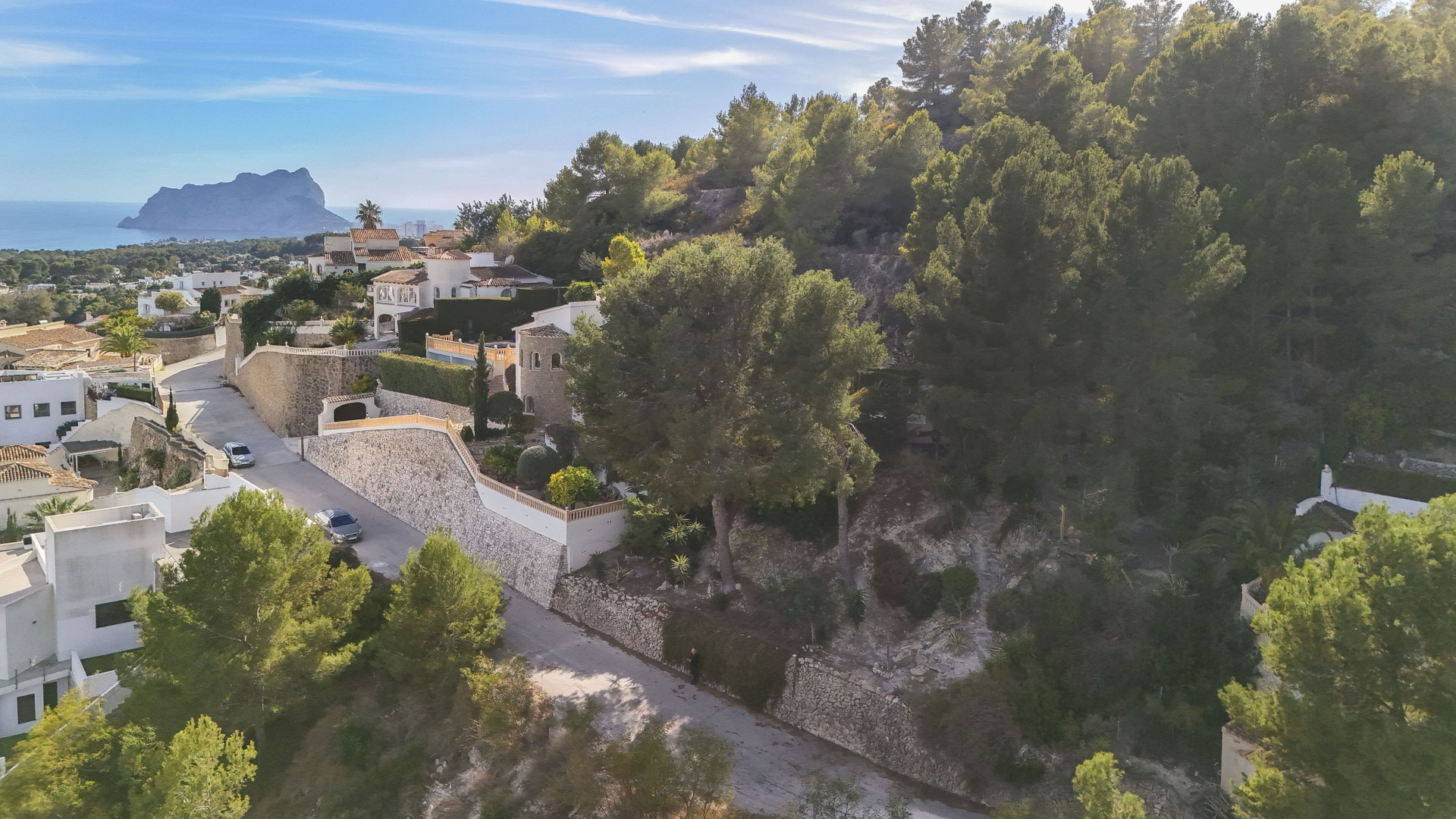 Luchtfoto van het hooggelegen perceel in Benissa, met de natuurlijke pijnbomen en een panoramisch uitzicht op zee met de Peñón de Ifach in de verte.