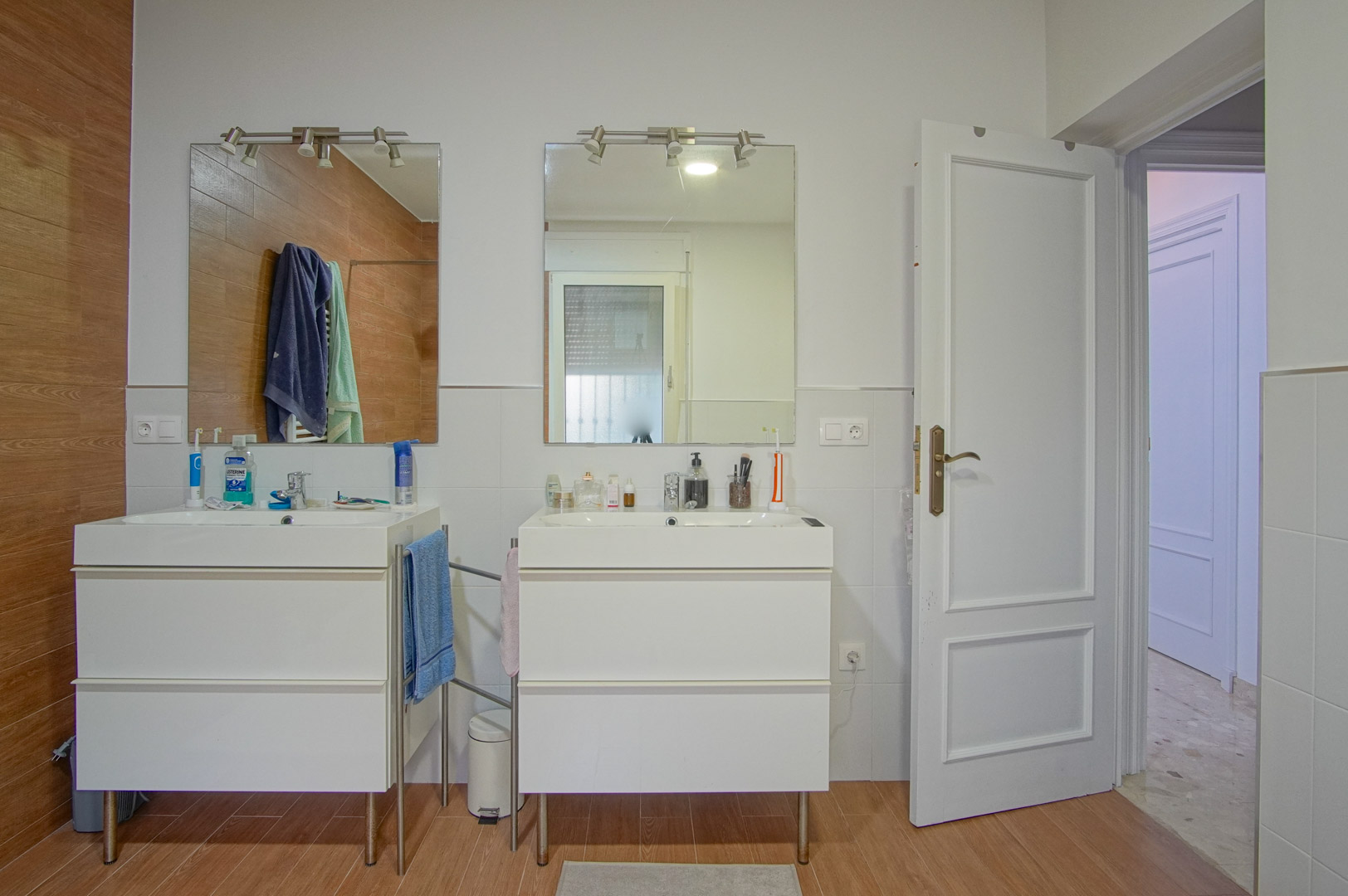 Badkamer met dubbele wastafel op afzonderlijke witte meubels, individuele spiegels met verlichting en wandafwerking in een mix van wit en houtlook.