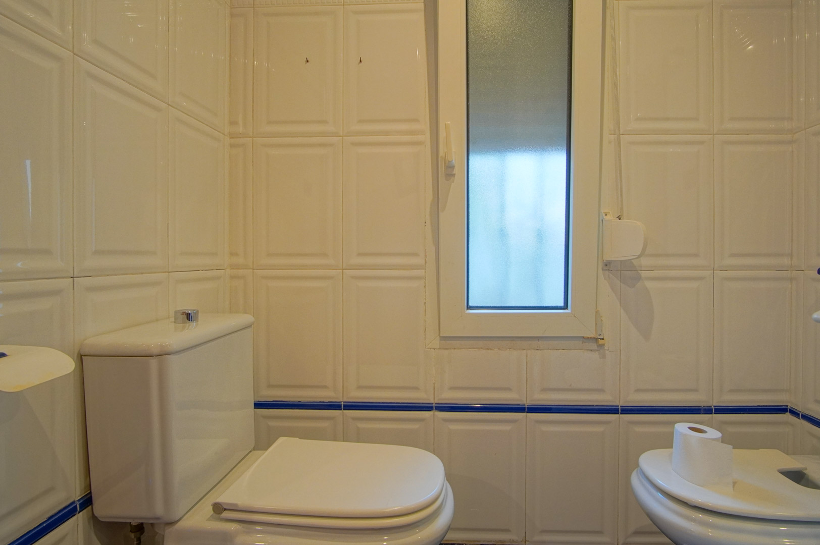 Badkamer met witte reliëftegels en een blauwe sierrand. Voorzien van een toilet, bidet en een raam met matglas voor natuurlijke lichtinval.
